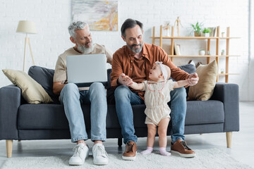 Smiling homosexual couple with laptop playing with toddler daughter at home