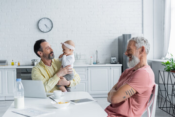 Homosexual freelancer holding baby daughter near cheerful partner in kitchen