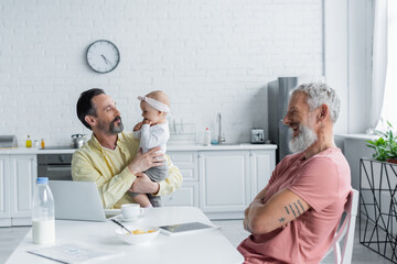 Naklejka premium Homosexual freelancer holding baby daughter near cheerful partner in kitchen