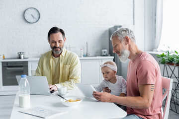 Homosexual man holding digital tablet near daughter and partner in kitchen