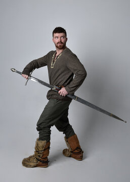 Full Length  Portrait Of  Young Handsome Man  Wearing  Medieval Celtic Adventurer Costume.  Standing Pose Holding A Sword, Isolated On Studio Background.