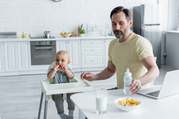 Mature man holding baby bottle near laptop, breakfast and daughter on high chair