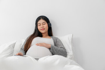 Asian happy pregnant woman is sitting on bed and and touching her belly and wear a headphone to listen the music. pregnancy, motherhood, people and expectation concept