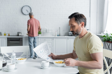 Homosexual man reading news near breakfast and blurred partner in kitchen