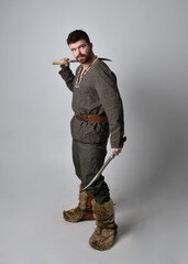 Full length  portrait of  young handsome man  wearing  medieval Celtic adventurer costume.  Standing pose holding a sword, isolated on studio background. - obrazy, fototapety, plakaty