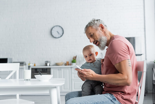 Man holding cellphone near toddler kid and breakfast in kitchen