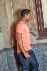 Dressing in a light orange short sleeve V neck shirt, jeans,  a young handsome black guy is standing by a window, interestedly looking down..