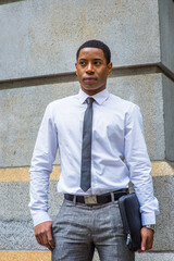 Dressing in white shirt, a black tie, gray pants, carrying a briefcase, a young handsome black college student is standing by a stone wall, confidently looking forward.