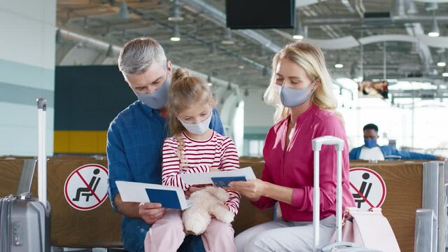 Portrait of the cheerful caucasian parents with cute daughter sitting in the airport with baggage and examining tickets while waiting for their flight to the vacation. People wearing face masks