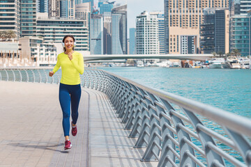 Happy woman wearing headphones and sunglasses trains and does running and fitness on the sidewalk of the embankment in the Dubai Marina area