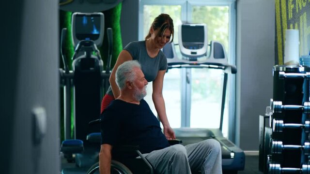 Old man in a wheelchair trains his shoulder in the gym. A female personal trainer helps a person with a disability to exercise