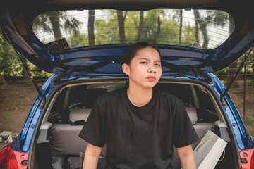 An unimpressed teenager sitting down on the open back of a hatchback car. An antsy tomboyish asian...