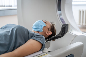 Obraz premium A woman in a medical mask lies on the tomograph table. woman is undergoing computed axial tomography examination in a modern hospital.