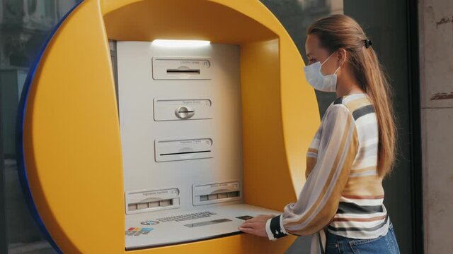 Woman wearing medical mask for prevent infection coronavirus using ATM machine to withdraw money on city street. Girl in bank terminal, tourist after quarantine is over, the end of lockdown. Barcelona