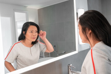Asian mature woman putting mascara looking in LED light frame mirror applying makeup getting ready...