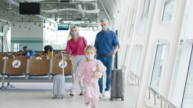Caucasian nice cheerful mother, father and their daughter wearing protective masks going through the passage in the airport with suitcases on the wheels to the plane as having vacations tour