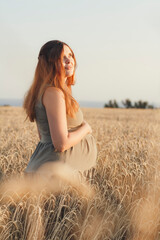 young pregnant woman walks on wheat field at sunset, expectant mother with relax in nature stroking her belly with hand, happy pregnancy concept