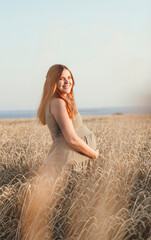 beautiful young pregnant woman walks on wheat field at sunset, expectant mother with relax in nature stroking her belly with hand, happy pregnancy concept