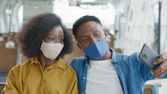 En Young afro american spouses wearing protective masks sitting in the airport and taking selfie photos to the camera of smartphone