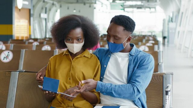 Happy multiracial couple wearing protective masks sitting in the airport with each other and looking at their passports, while waiting at the departure