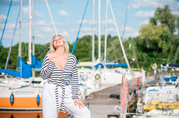 Pretty blonde woman of plus size, American or European appearance near yachts, enjoying life. Young lady with excess weight, stylishly dressed at striped jumper and white pants .Natural beauty