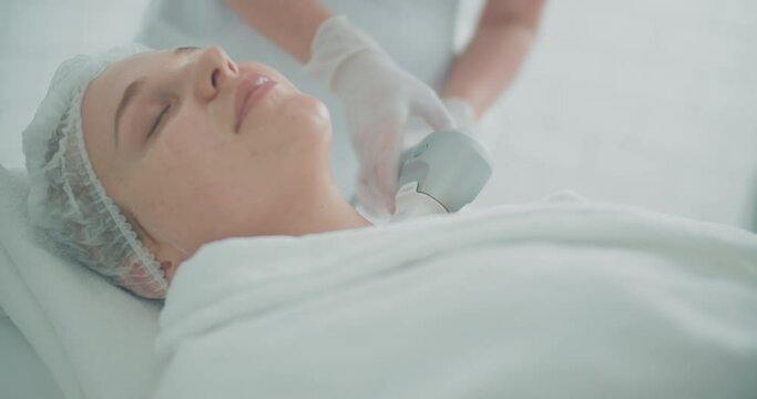 A close-up portrait of a female client, she is laying with closed eyes in a bathrobe and disposable hat while having a SMAS lifting procedure by using the Ultraformer apparatus. 