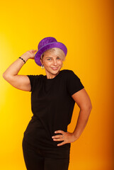 mature woman in a joyful mood, the emotion of victory, on a yellow background in a purple hat shows her super thumb, holds on to the hat. place for text.