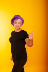 Fototapeta na wymiar mature woman in a joyful mood, the emotion of victory, on a yellow background in a purple hat shows her super thumb, holds on to the hat. place for text.