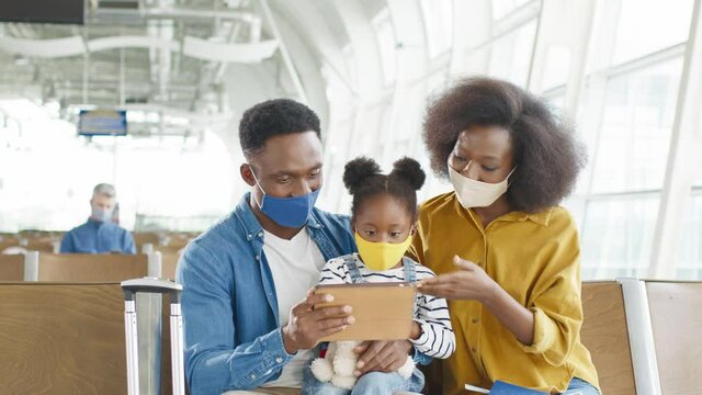 Multiracial family with a daughter wearing protective masks sitting in the departure hall at the airport, talking and watching something on the tablet computer