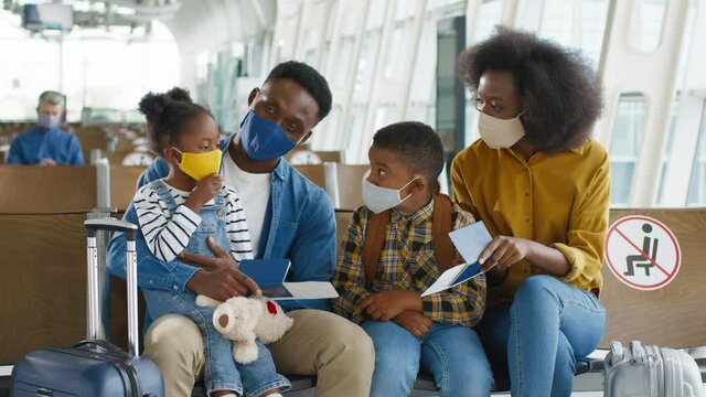 Portrait of the cheerful multiracial parents with cute kids sitting in the airport with baggage and smiling to each other while waiting for their flight to the vacation. People wearing face masks
