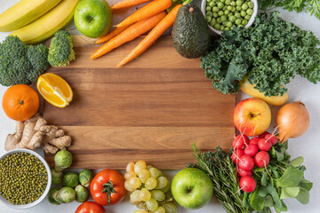 Variety of raw green vegetables salads, carrots, tomatoes, bananas, peas, corn, broccoli, savoy cabbage as frame round empty white chopping board. Food background. Top view, space for text