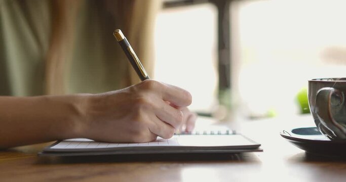 Close up of woman's hand writing down on paper notebook with coffee cup on table sitting in cafe