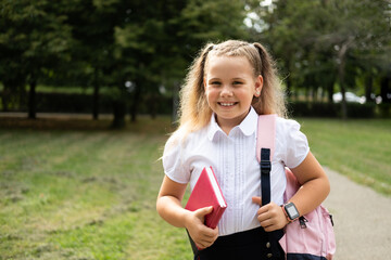 blonde smiling schoolgirl in school uniform holding notebook with pink backpack back to school...
