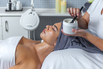 Beautician spreading cream from the container to apply on face of client woman in the skin care treatment in spa salon