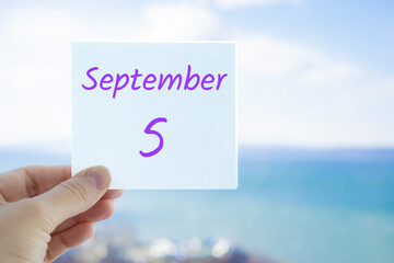 5th September. Hand holding sticker with text September 5 on the blurred background of the sea and sky. Copy space for text. Month in calendar concept