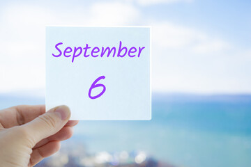 6th September. Hand holding sticker with text September 6 on the blurred background of the sea and sky. Copy space for text. Month in calendar concept