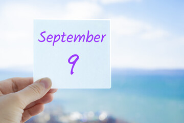 9th September. Hand holding sticker with text September 9 on the blurred background of the sea and sky. Copy space for text. Month in calendar concept