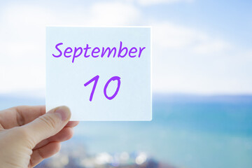 10th September. Hand holding sticker with text September 10 on the blurred background of the sea and sky. Copy space for text. Month in calendar concept