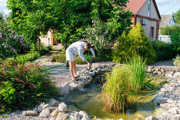 Young caucasian man cleans artificial garden pond bottom with high-pressure washer nozzle from mud and sludge. Spring and summer pond care work