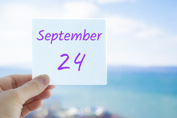 24th September. Hand holding sticker with text September 24 on the blurred background of the sea and sky. Copy space for text. Month in calendar concept