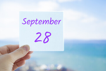 28th September. Hand holding sticker with text September 28 on the blurred background of the sea and sky. Copy space for text. Month in calendar concept