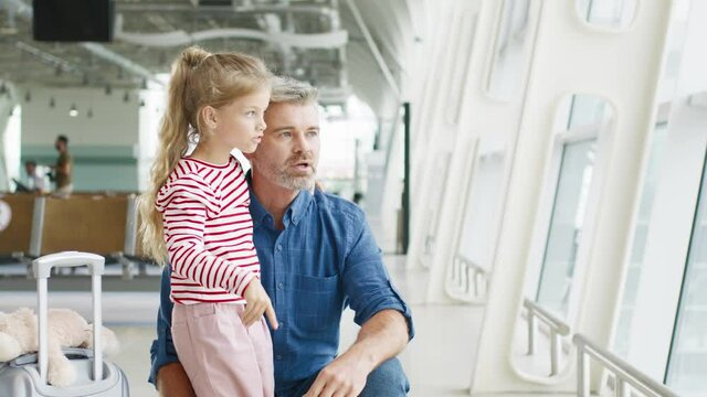 Senior father and his lovely daughter looking at airplanes through the glass window and chatting with pleasure emotions. Travel with baby at the airport concept