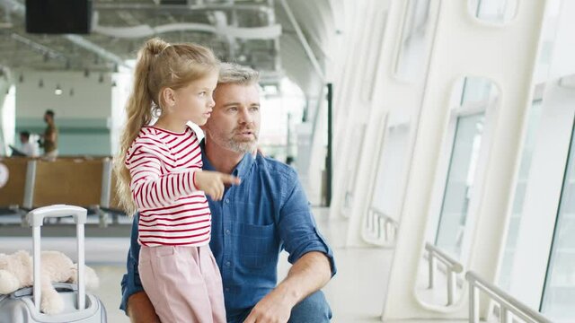 Senior father and his lovely daughter looking at airplanes through the glass window and chatting with pleasure emotions. Travel with baby at the airport concept
