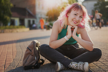 Stylish young smiling woman with multicolored pink hair sitting on the pavement in a sunny city holding hands in the shape of a heart