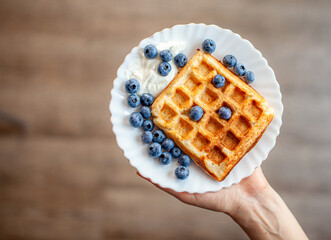 A woman holds in her hand a plate on which fresh waffles with berries are ready. Homemade waffles...