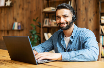 Smiling Indian male freelancer wearing headset and smart casual wear using laptop for connection...