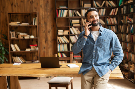 Happy Inspired Indian Male Small Business Owner Talks On The Smartphone Standing In Modern Loft Office, Smiling Successful Contemporary Man Manager In Casual Wear Holding Phone Call, Copy Space