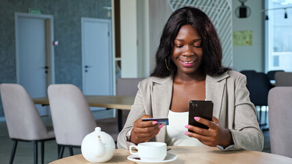 Cheerful black woman in stylish jacket pays online with credit card and mobile phone sitting at...