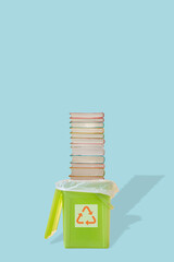 Cover page with green garbage trash bin with bag and a pile of paperback books with its shadow and recycling symbol at blue solid background with copy space. Concept of paper recycling.