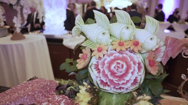 Meat assortment on the festive buffet table. The banquet is decorated with carved fish and a watermelon flower. A product of art. Handmade. Fruit carving. Carved figures